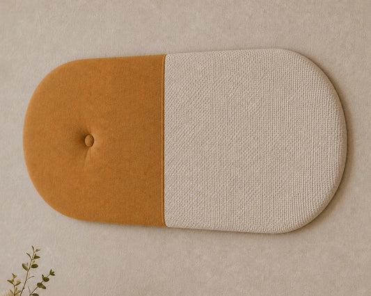 Close-up angled view of a two-tone upholstered wall panel, highlighting its mustard fabric, textured cream section, rounded shape, central seam and button detail